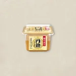 Plastic tub of miso paste with a yellow label and red vertical text, placed on a light beige surface.