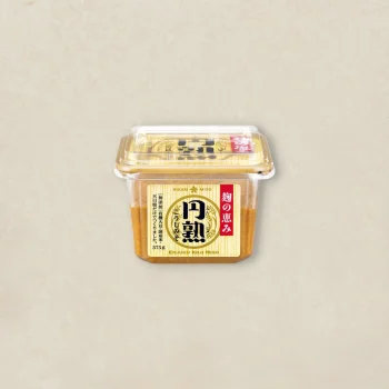 Plastic tub of miso paste with a yellow label and red vertical text, placed on a light beige surface.