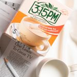 Box of 3:15PM Original Milk Tea with a cup illustration on orange-white packaging, on a newspaper with pencils and mugs nearby