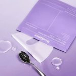 Lavender background with a purple facial mask package, a white sheet mask, and a spoonful of charcoal powder with small liquid droplets.