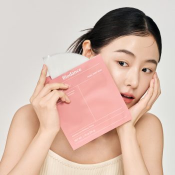 Close-up of a woman holding a pink Biodance facial sheet mask against her cheek.
