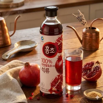 Bottle of 100% pomegranate juice with sliced pomegranate images beside a glass of red juice on a wooden table, with pomegranate fruit nearby and copper kitchenware in the background
