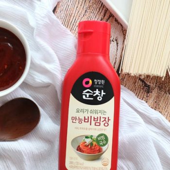 Red bottle of Korean bibimbap sauce on a wooden table with a spoon and noodles nearby; label shows 만능비빔장 and 순창 badge.