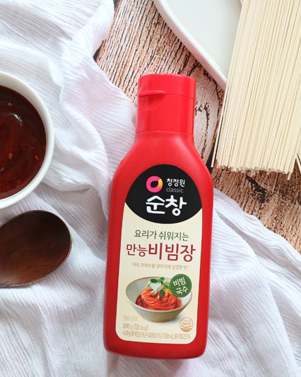 CHUNGJUNGONE Bibimjang Red bottle of Korean bibimbap sauce on a wooden table with a spoon and noodles nearby; label shows 만능비빔장 and 순창 badge.