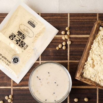 Top-down view of a diagonal powder package, a bowl of creamy sauce with sesame seeds, and a wooden scoop of flour on a dark wooden board with scattered soybeans and a green plant nearby.